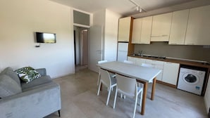Een modern appartement met een grijze bank, een eettafel met stoelen, een keuken met witte kasten en een wasmachine.