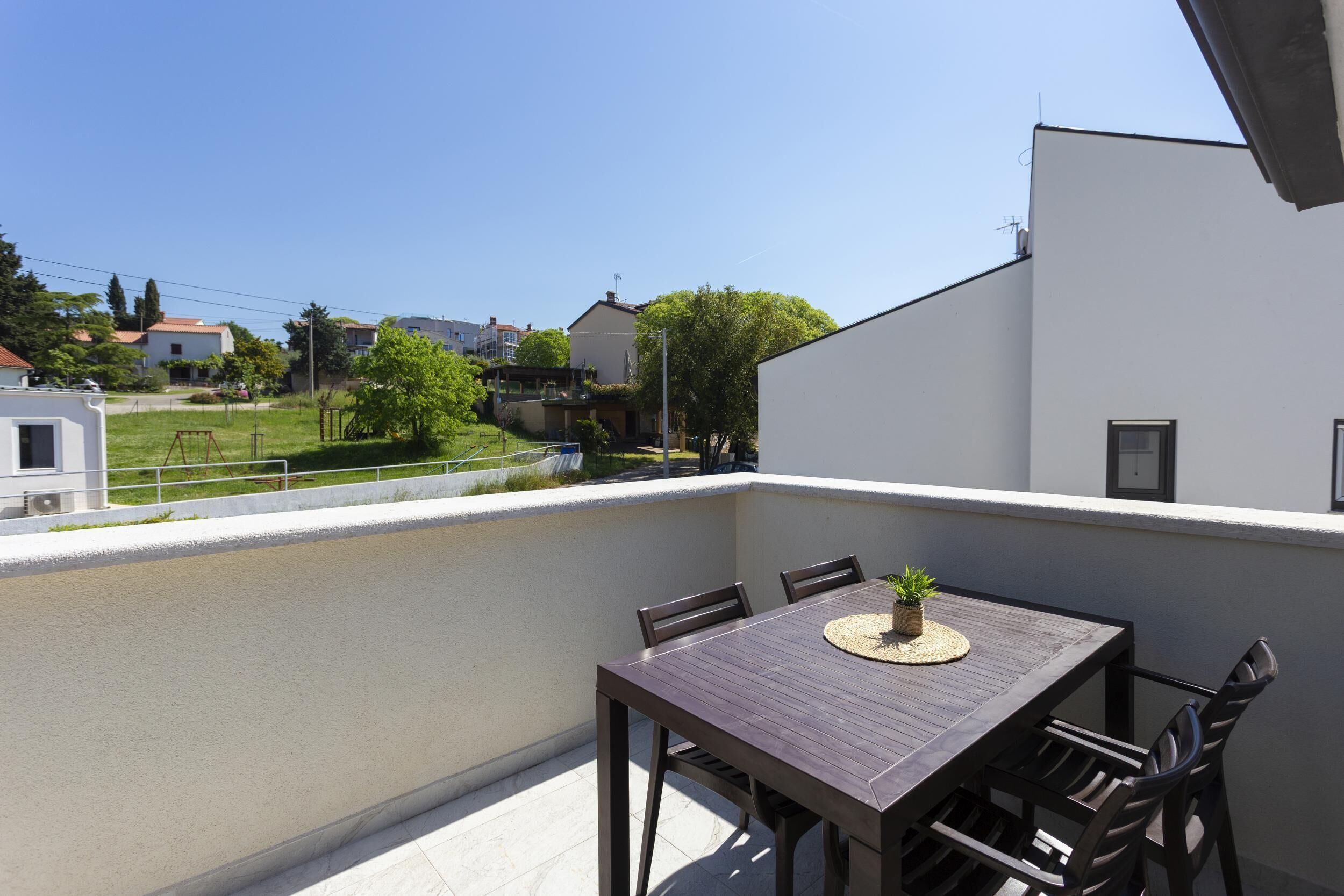 Ein Balkon mit Holztisch und -stühlen, einem kleinen Topfpflanzen und Blick auf ein Wohngebiet mit Häusern und Grünflächen.