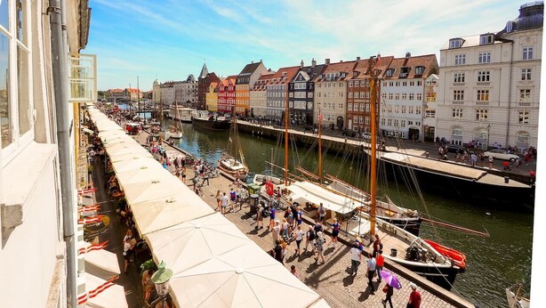A riverside promenade with a row of colorful buildings, a dock with boats, and people walking along the path. - Charming Nyhavn Apartment - Stunning Canal Views (København)
