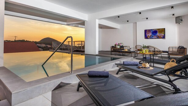 Piscina infinita en la azotea con sillones de descanso y una mesa, con vistas a un paisaje urbano al atardecer.