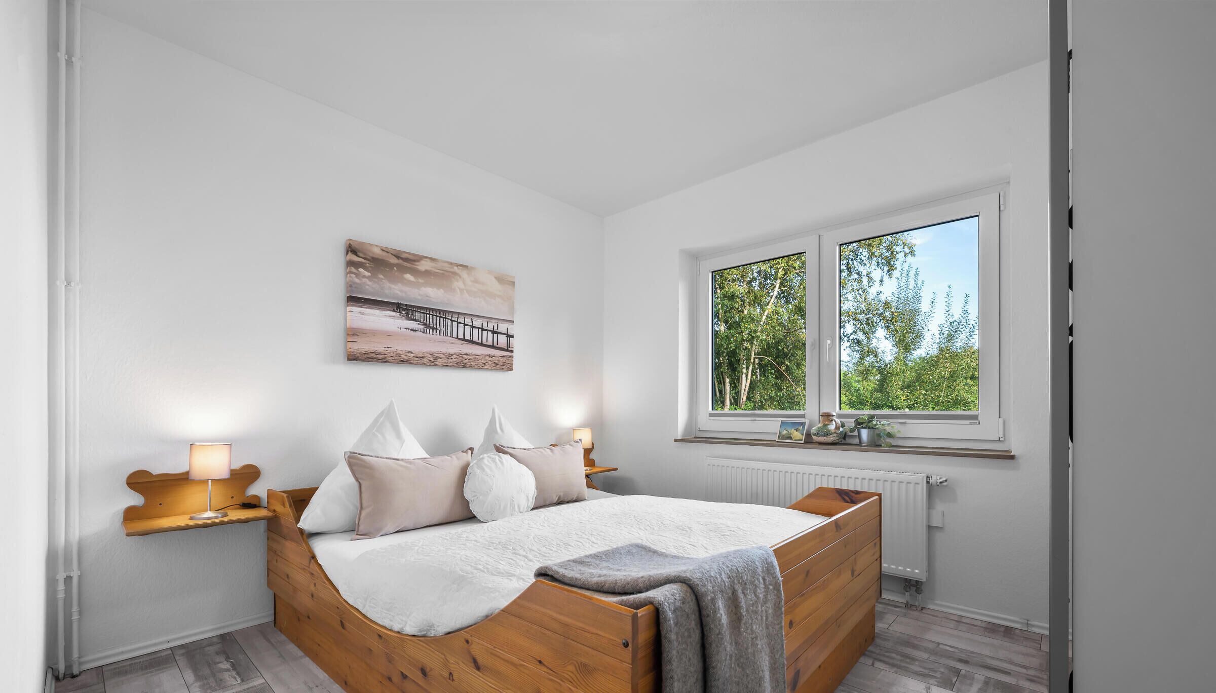 A bedroom with a wooden bed, white walls, a window with a view of greenery, and a painting on the wall.