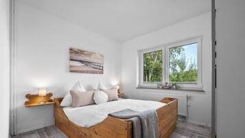 A bedroom with a wooden bed, white walls, a window with a view of greenery, and a painting on the wall.