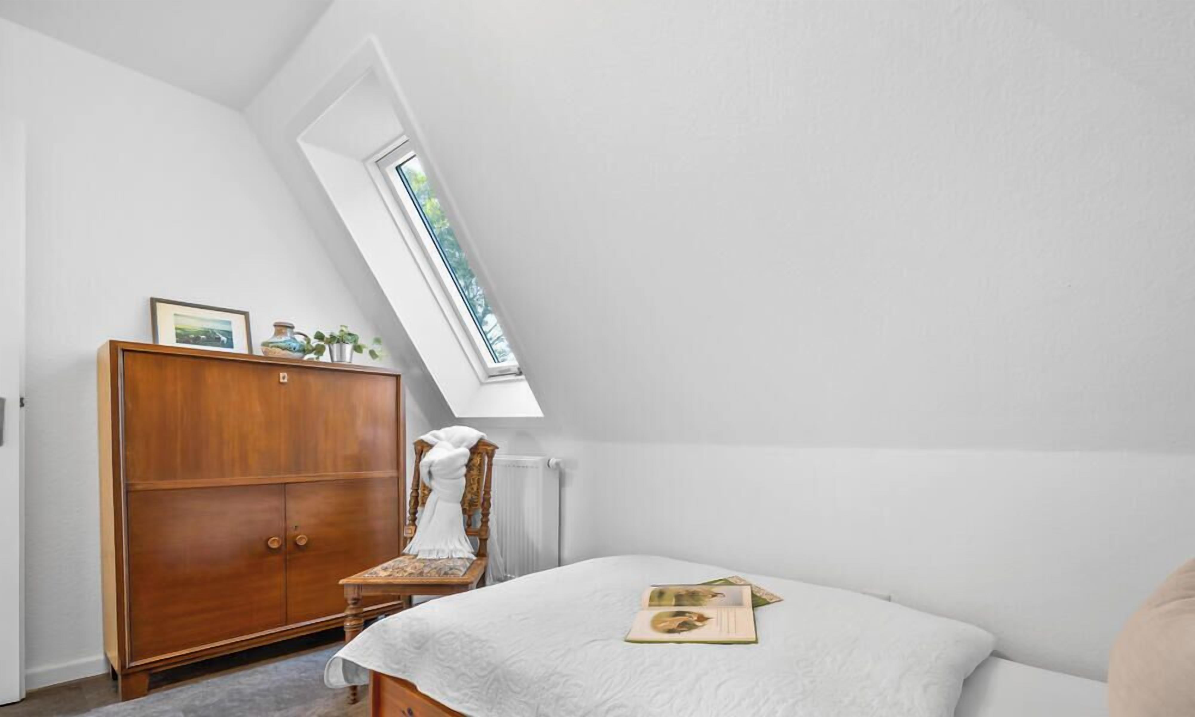 Un dormitorio con techo inclinado, un armario de madera, una cama con colcha y una ventana con vistas a árboles.