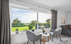 Ein modernes Wohnzimmer mit großem Fenster, einem grauen Sofa, zwei grauen Sesseln und einem kleinen Tisch mit Blumentopf.
