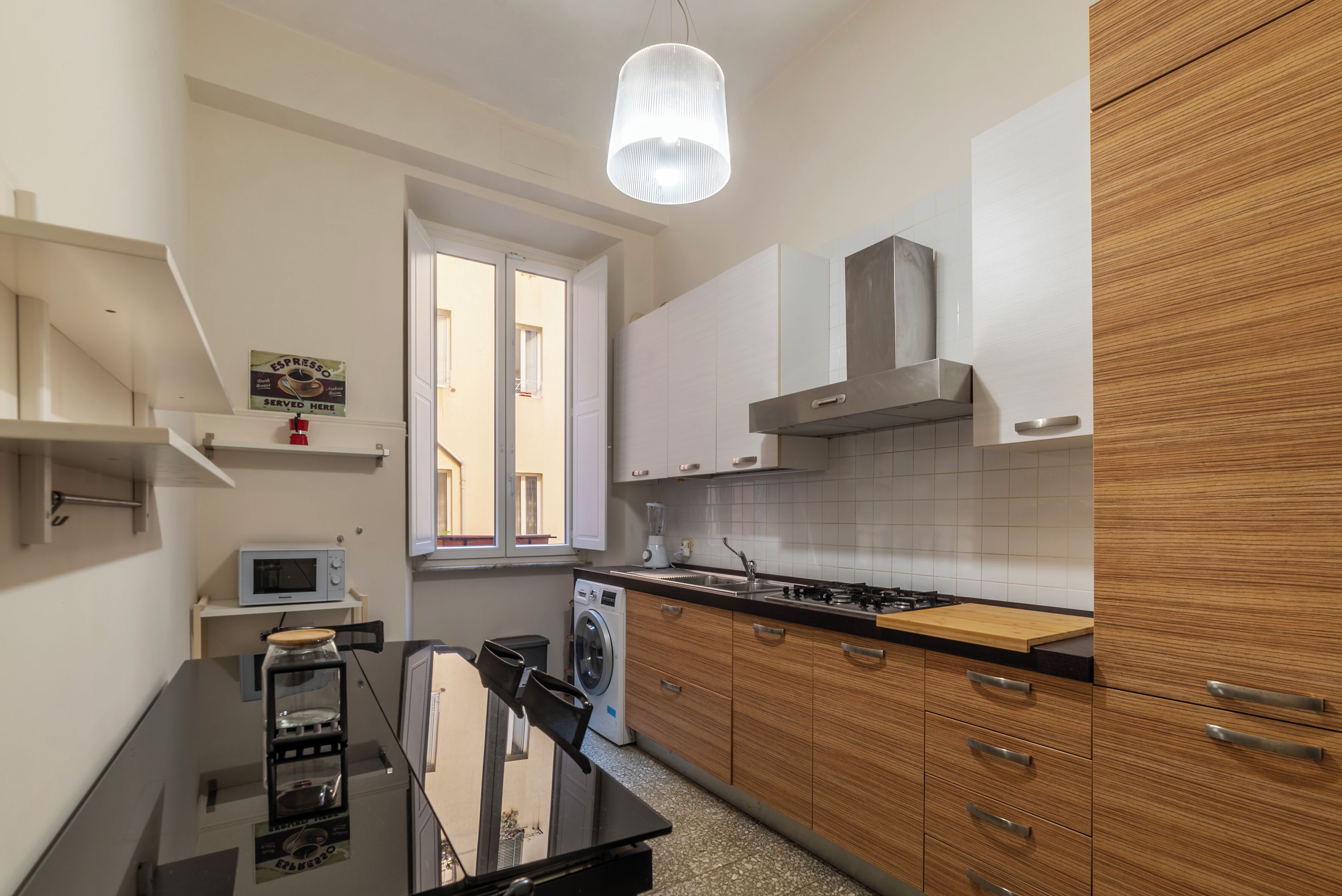 A compact kitchen with a microwave, washing machine, and a stove. There is a window providing natural light and a hanging light fixture.