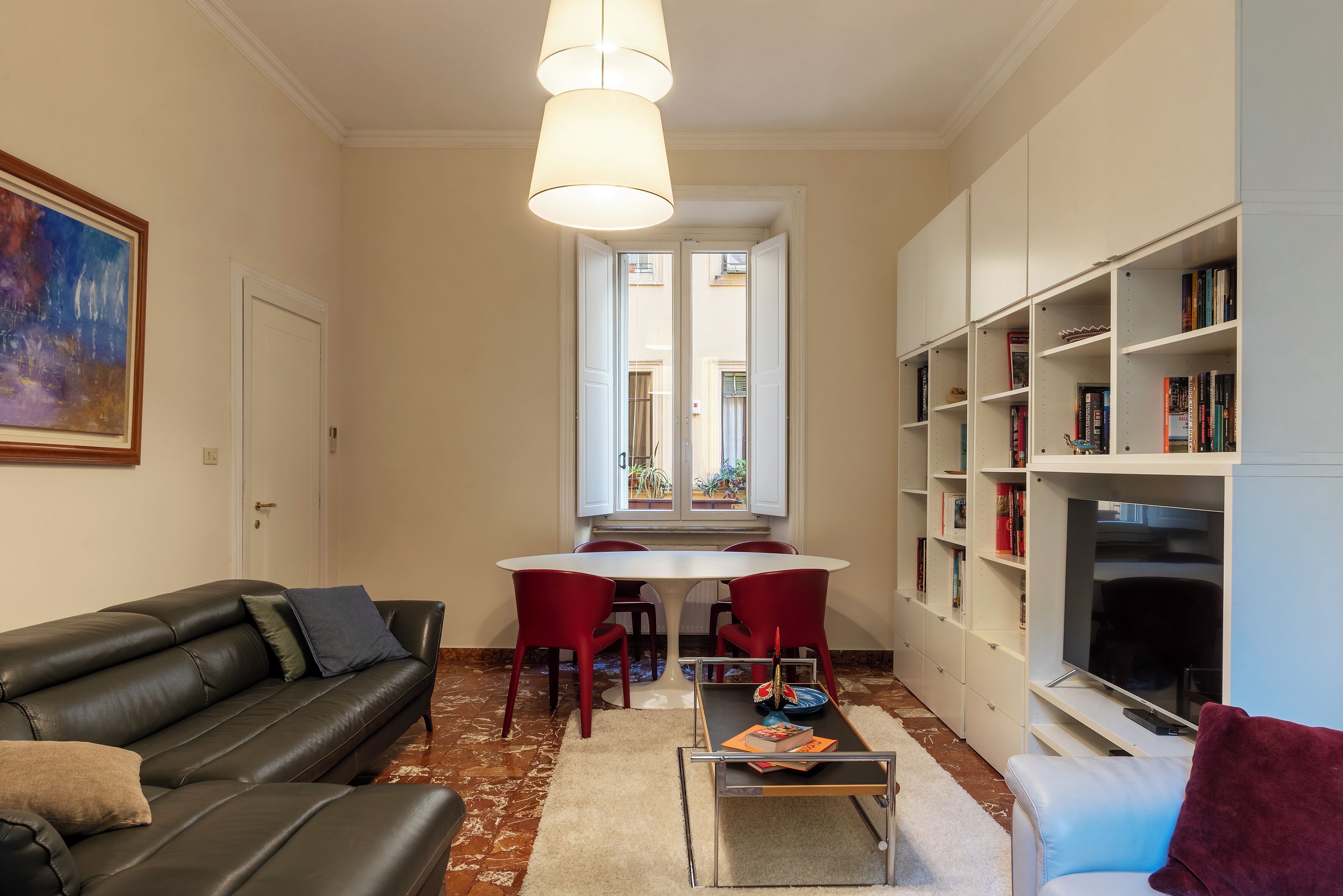 Una sala de estar con un sofá de cuero negro, una mesa de centro, una mesa de comedor con sillas rojas, una estantería y un televisor.