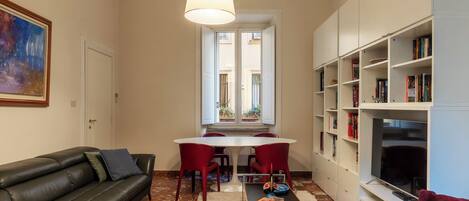 Una sala de estar con un sofá de cuero negro, una mesa de centro, una mesa de comedor con sillas rojas, una estantería y un televisor.