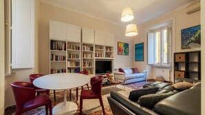 Ein modernes Wohnzimmer mit einem weißen Esstisch, roten Stühlen, einem schwarzen Ledersofa und einem Bücherregal voller Bücher.