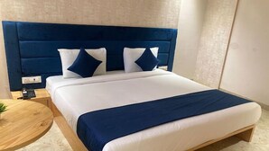 A neatly made bed with a blue headboard and white bedding, a round wooden side table, and a small plant. - JMC HOTEL STAR INN (Indore)