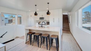 Une cuisine moderne avec des meubles blancs, un îlot central doté de tabourets, un éclairage suspendu et une vue sur l’extérieur.