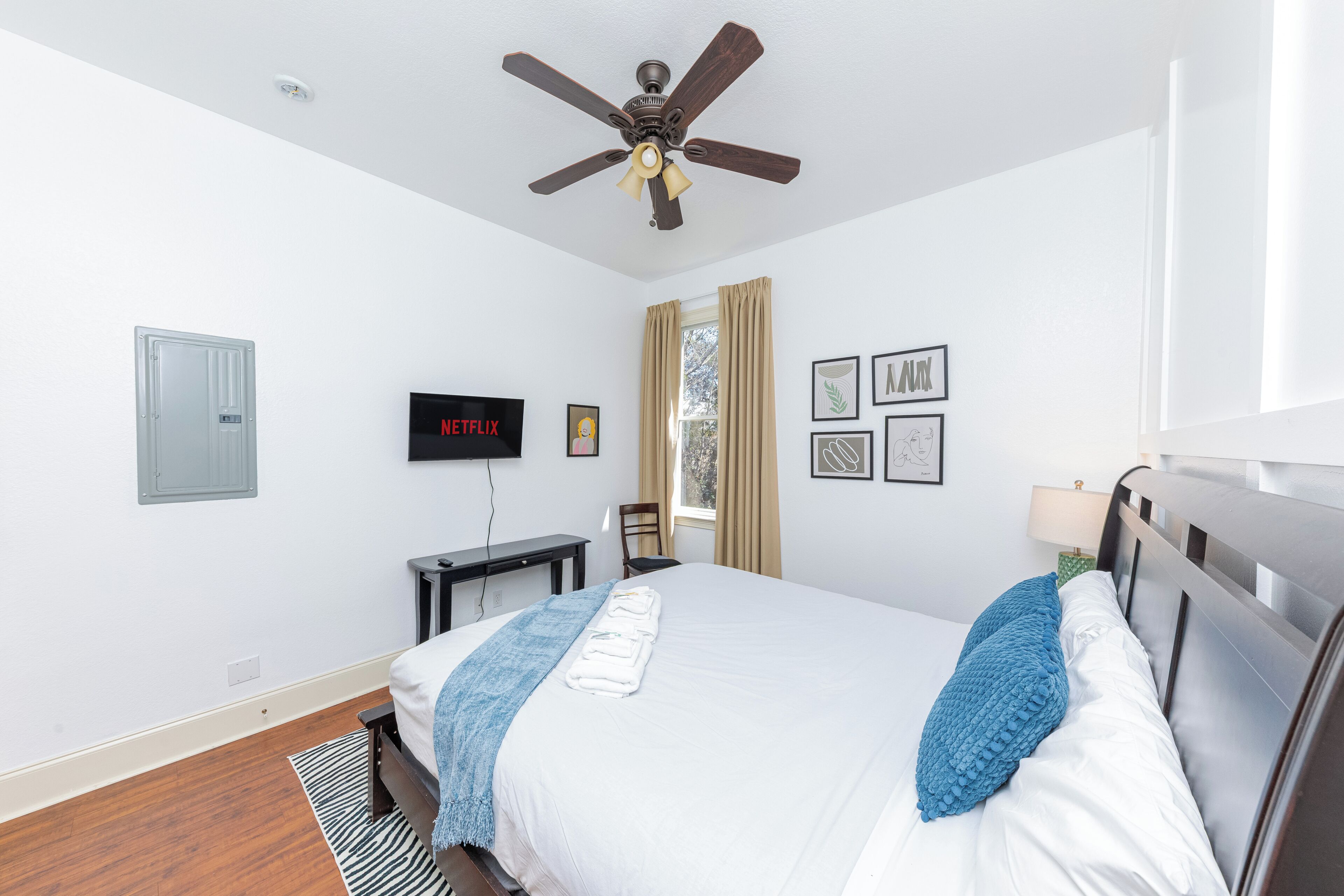 A hotel room with a bed, a ceiling fan, a TV, a desk, and a window with curtains.