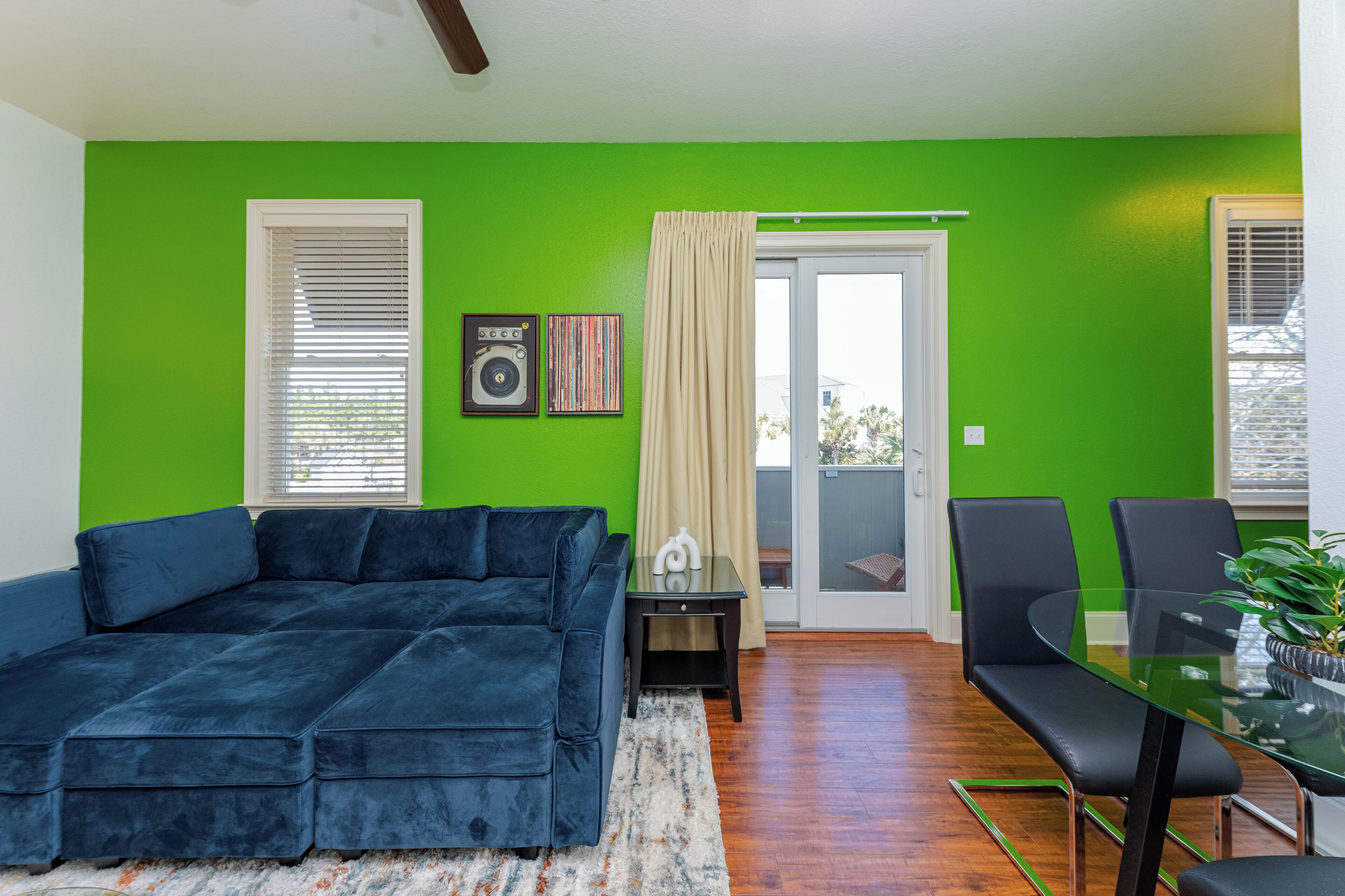 A living room with a blue sectional sofa, a glass dining table with chairs, a small side table, and a green wall.
