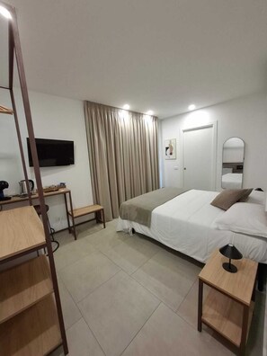Quarto de hotel com cama, escrivaninha com cadeira, televisão fixada na parede, espelho e janela com cortinas.