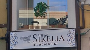 Ein zweistöckiges Gebäude mit einem Schild, auf dem "ROOMS SIKELIA" und eine Kontaktnummer stehen. Im Erdgeschoss befindet sich ein Blumentopf, und im Obergeschoss ein Balkon mit Stuhl und Tisch.