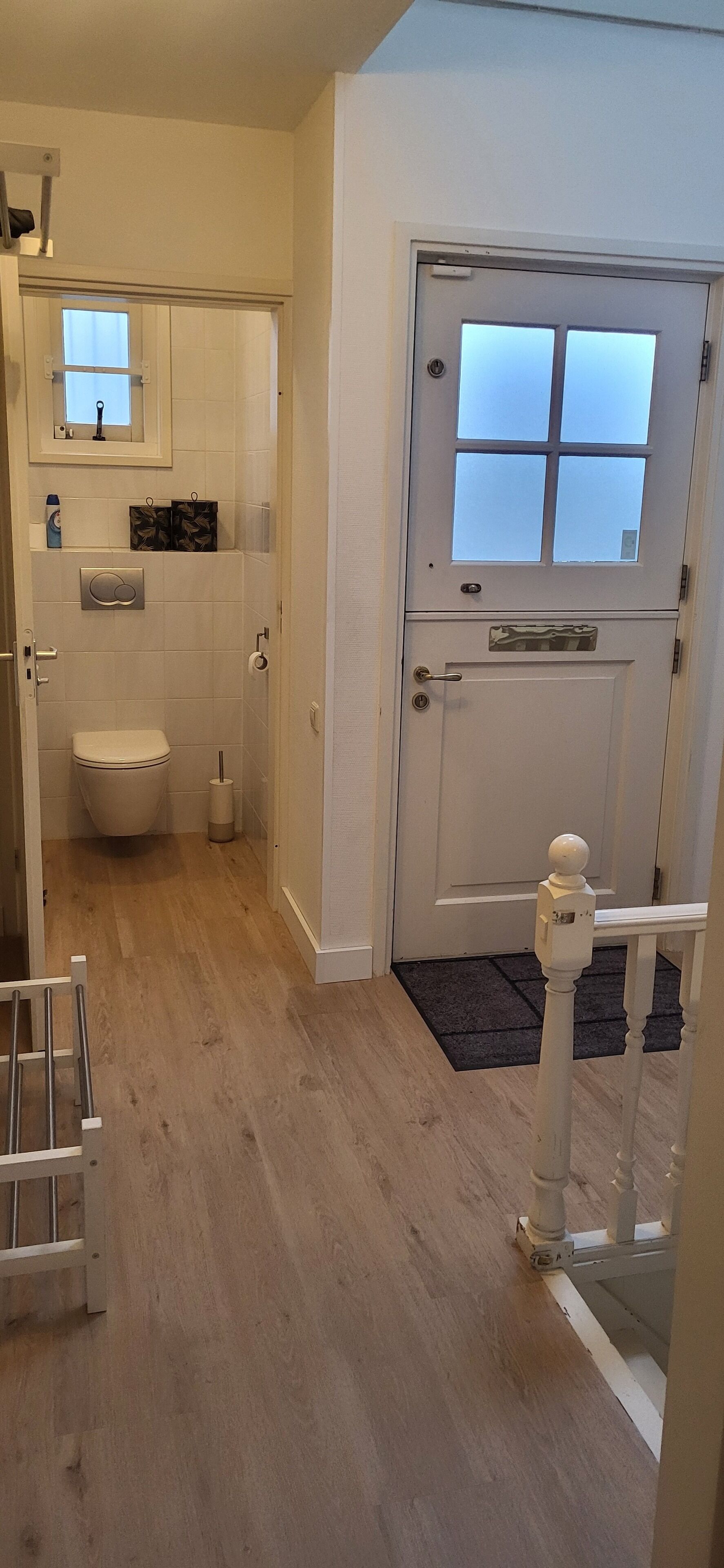 Un baño con inodoro, lavabo y espejo. Hay una puerta con ventanal y felpudo.