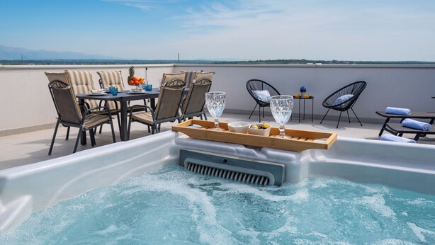 A rooftop terrace with a hot tub, dining table, chairs, and a view of the landscape.