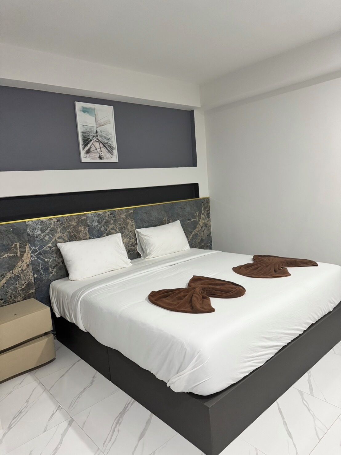 A modern hotel room with a large bed, a headboard with a marble design, a bedside table, and a framed picture on the wall.