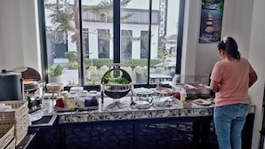 A breakfast buffet setup with various food items and condiments, a large window with a view of palm trees and buildings, and a person preparing food.
