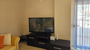 A living room with a flat-screen TV on a black stand, a yellow sofa with cushions, a small table, and a window with sheer curtains. - Albania Dream Holidays Apartment (Sarandë)