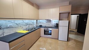 A compact kitchen with beige cabinets, a marble backsplash, a black countertop, a built-in oven, and a white refrigerator. - Albania Dream Holidays Apartment (Sarandë)