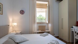 Ein Schlafzimmer mit Bett, Nachttisch mit Lampe, Fenster mit Blick, Heizkörper und Kleiderschrank.