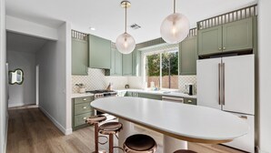 Een moderne keuken met groene kastjes, een groot eiland met krukken, hanglampen en uitzicht door het raam.
