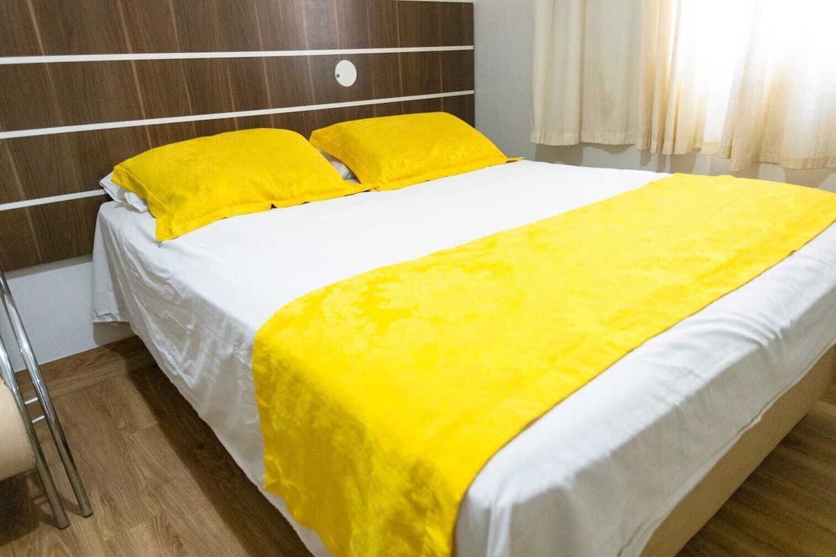 A bed with a white mattress and two yellow pillows, a wooden headboard, and a white curtain in the background.