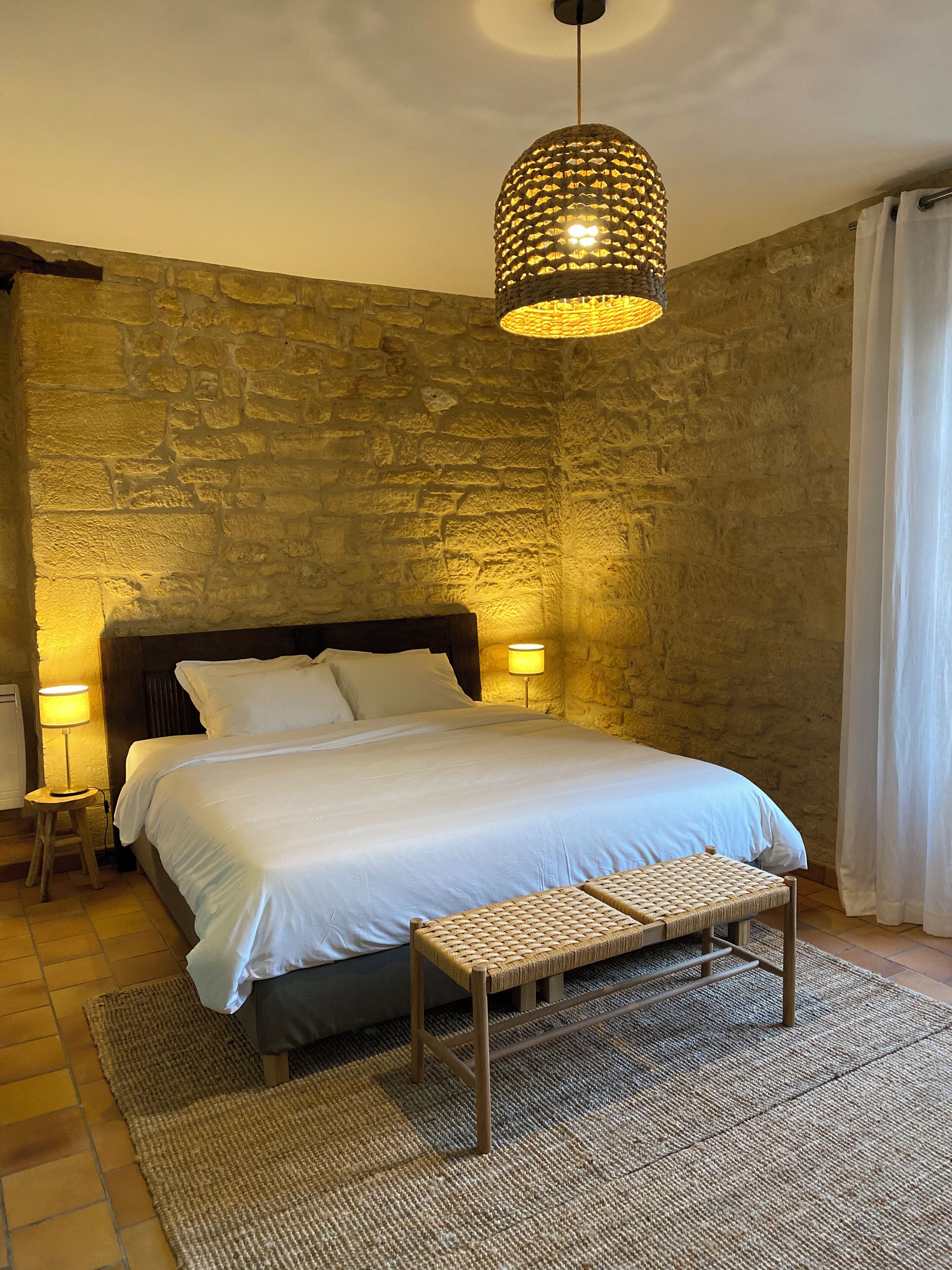 Un dormitorio con pared de piedra, una cama con sábanas blancas, dos lámparas de noche y un banco de fibras.