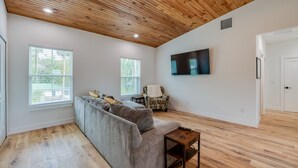 A spacious living room with a wooden ceiling, a grey sectional sofa, a patterned armchair, and a flat-screen TV mounted on the wall. - Forget your worries in this serene space. New 4-bedroom 4 bath house in Astor (Astor)
