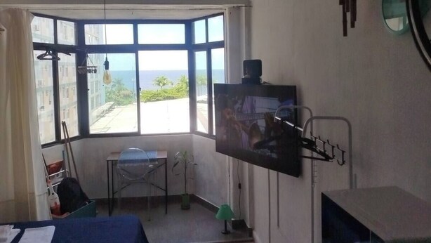 A room with a bed, a desk, a TV mounted on the wall, a window with a view of the sea, and a small table with a plant. - Sea view on the beach avenue - Embaré - Santos (Embaré Santos)