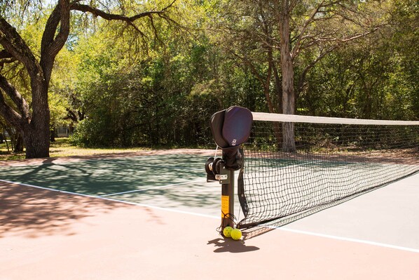 A tennis court with a net, trees, and a tennis ball rack.