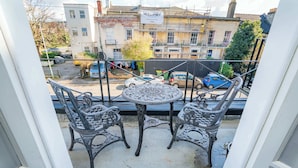 Ein Balkon mit kunstvollen Metallstühlen und einem kleinen Tisch, mit Blick auf eine Straße mit parkenden Autos und Gebäuden.