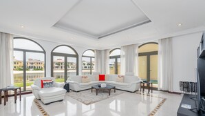 Een ruime woonkamer met een witte hoekbank, een salontafel en een flatscreen televisie. Grote boogvensters bieden uitzicht op een zwembad en gebouwen.