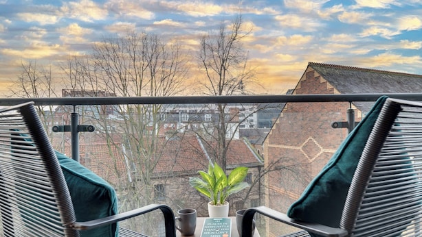 En balkon med to fletstole, et lille bord med en urtepotte og en bog, og udsigt til bygninger og træer under en overskyet himmel.