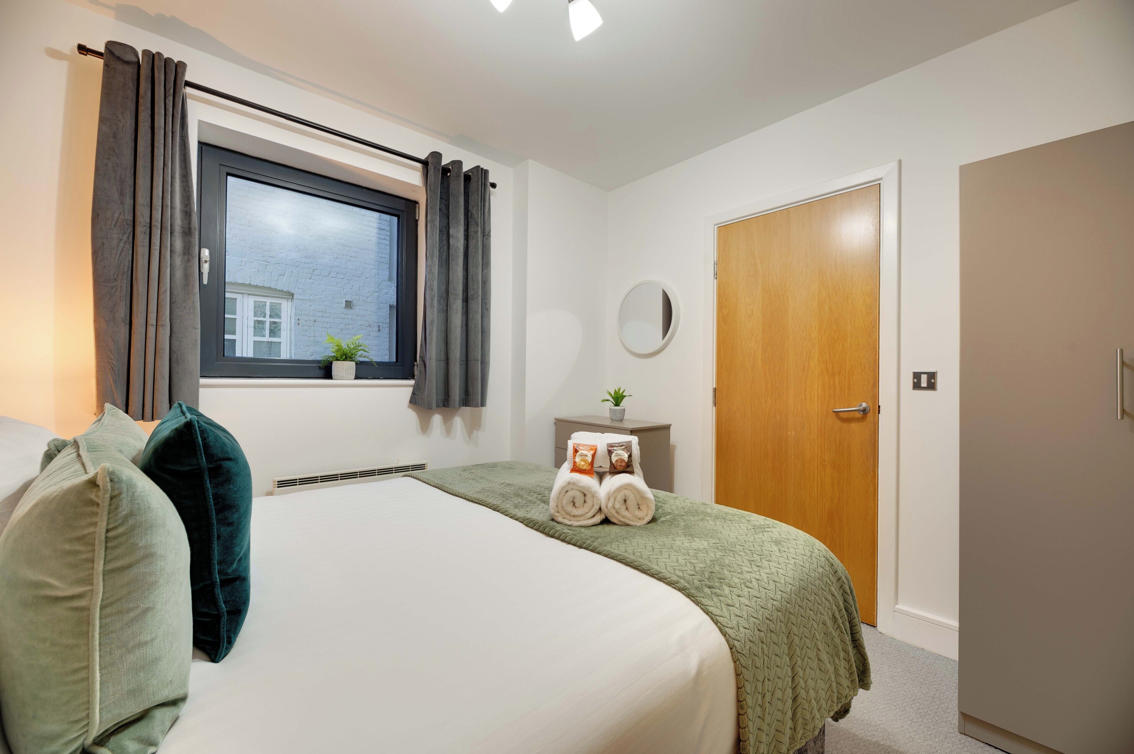 A hotel room with a large bed, a window with curtains, a bedside table with a plant, and a wooden door.