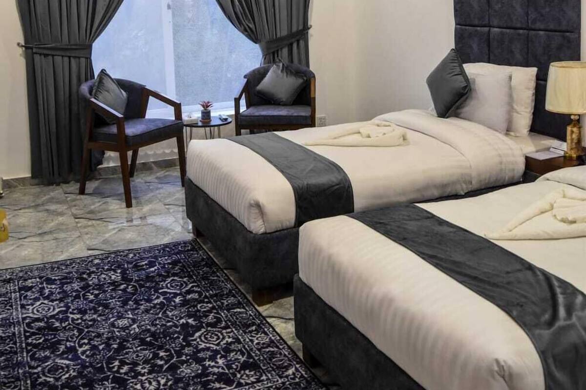 A hotel room with two beds, a ceiling fan, an air conditioner, a window with curtains, a small table, two chairs, and a patterned rug.