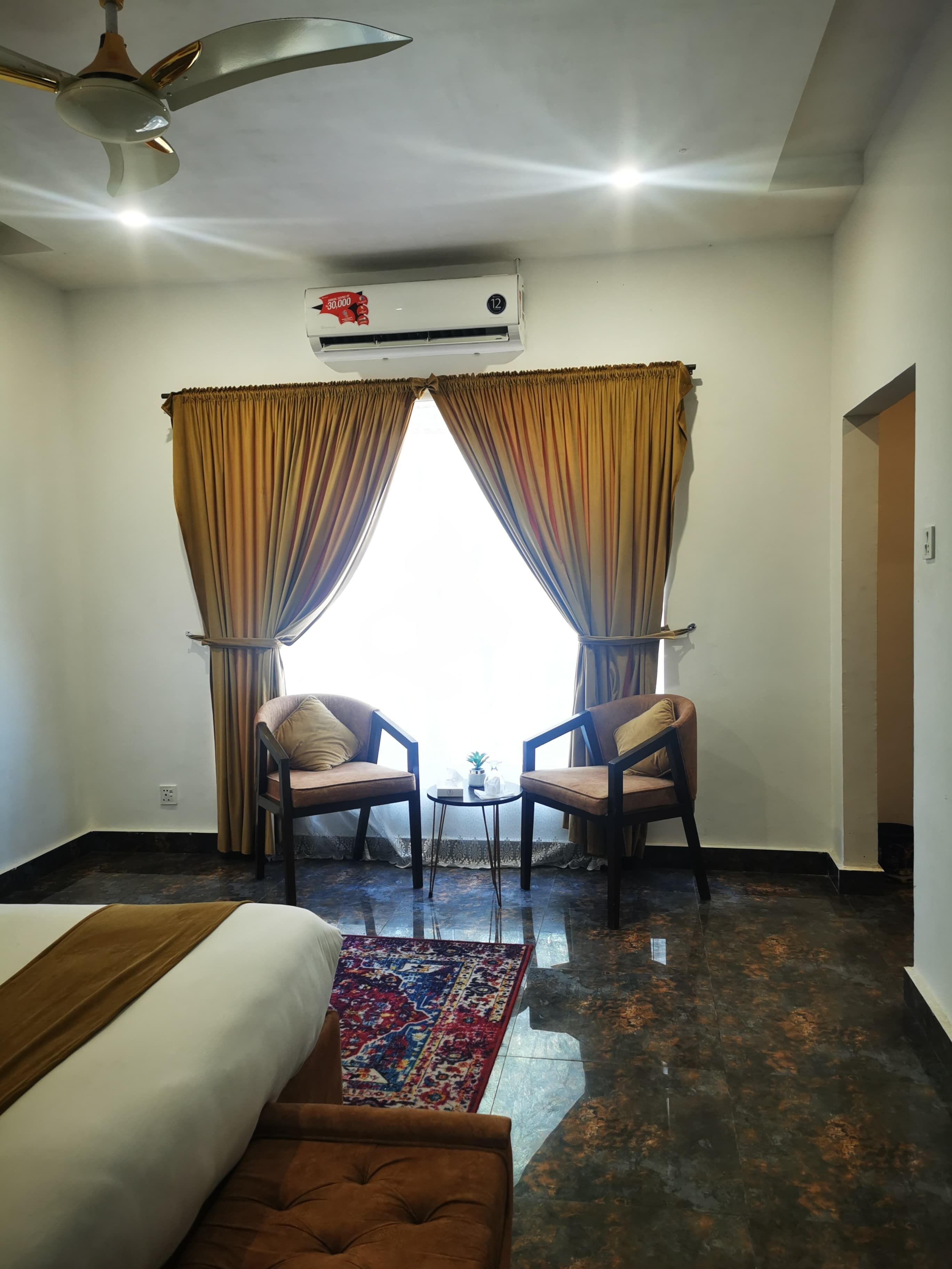 A hotel room with a bed, two armchairs, a small table, a ceiling fan, an air conditioning unit, and a patterned rug.