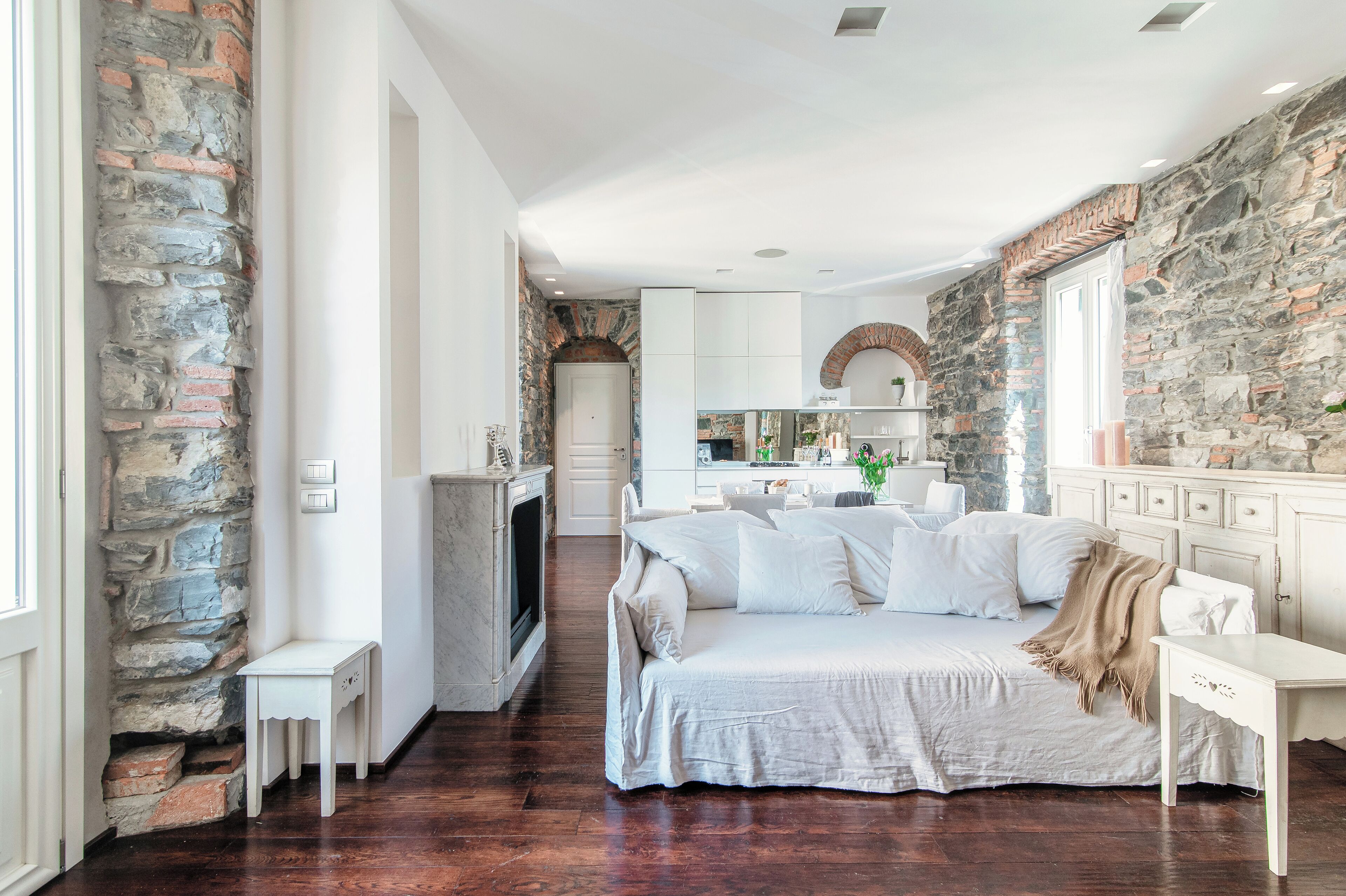 A spacious bedroom with a large bed, a fireplace, stone walls, and a view into the kitchen.