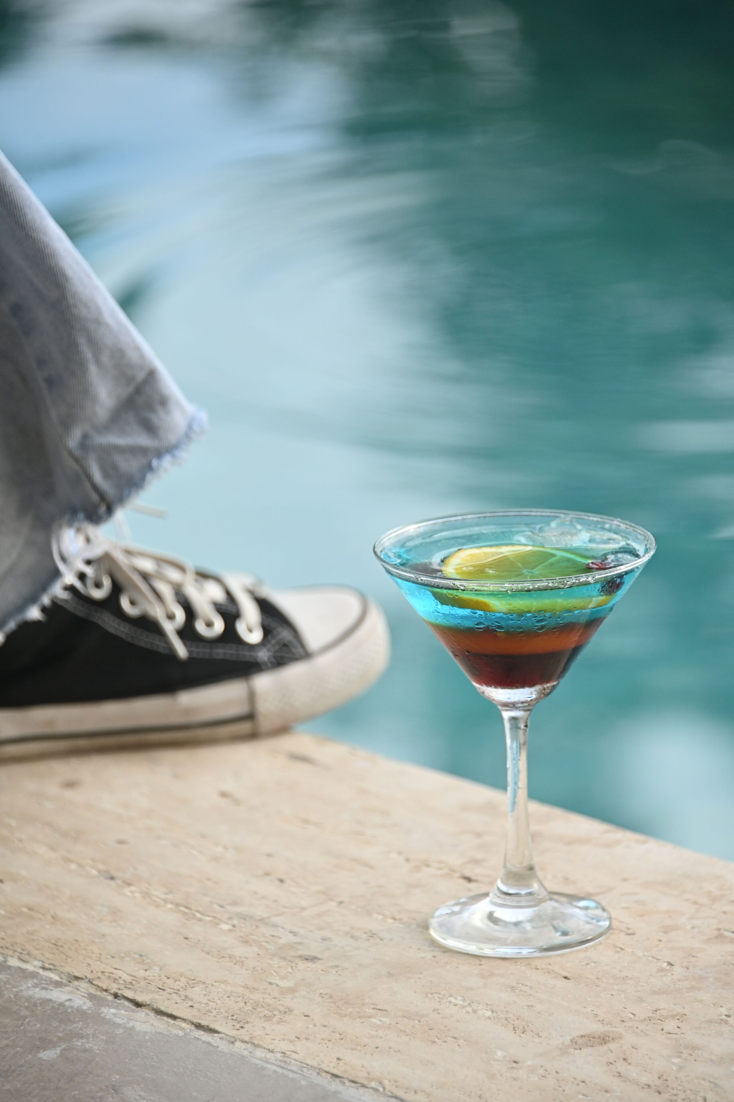 A martini glass with a lime slice on a concrete surface near a pool.