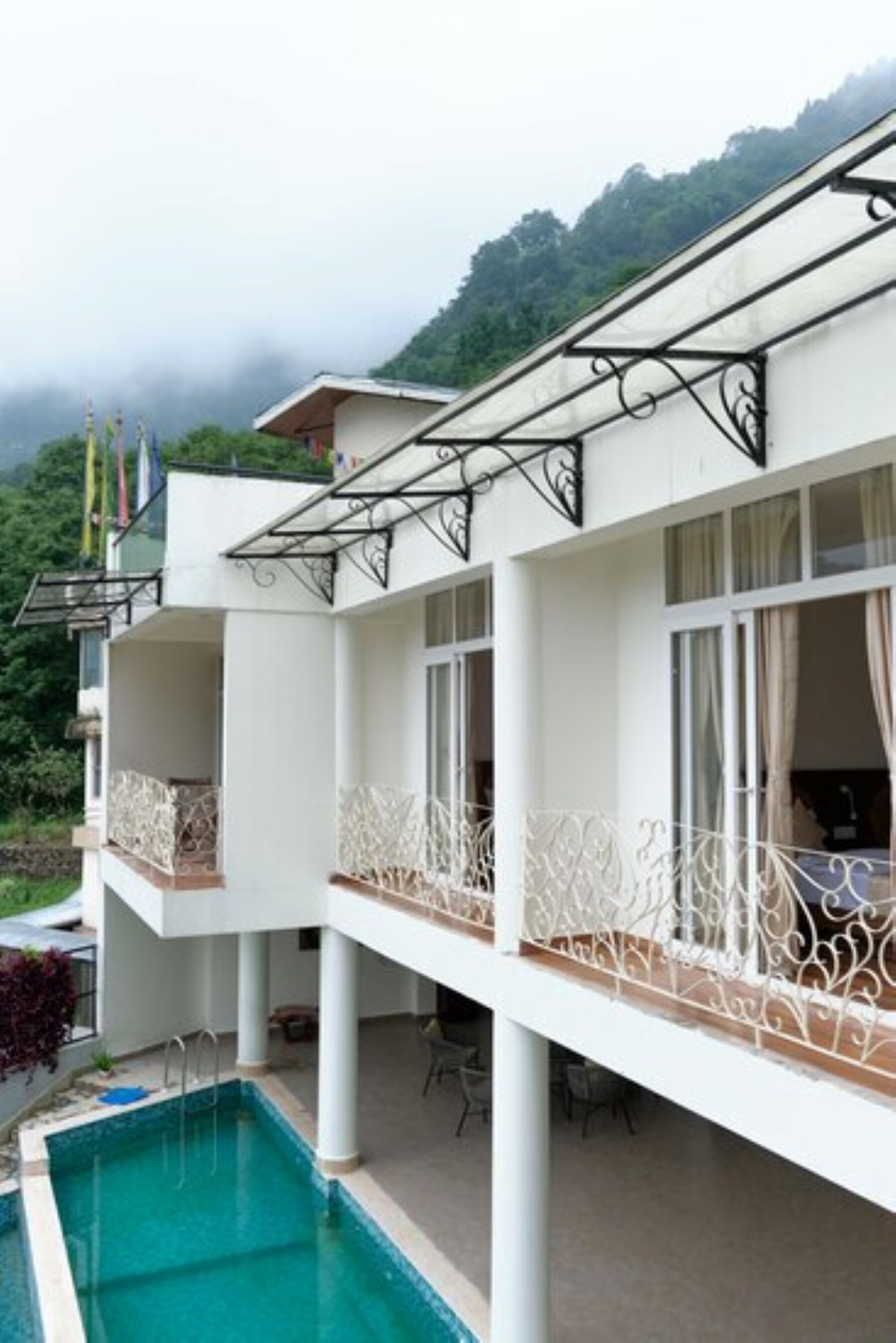 Un hotel moderno con piscina, balcones con barandillas blancas y vistas a la vegetación circundante.
