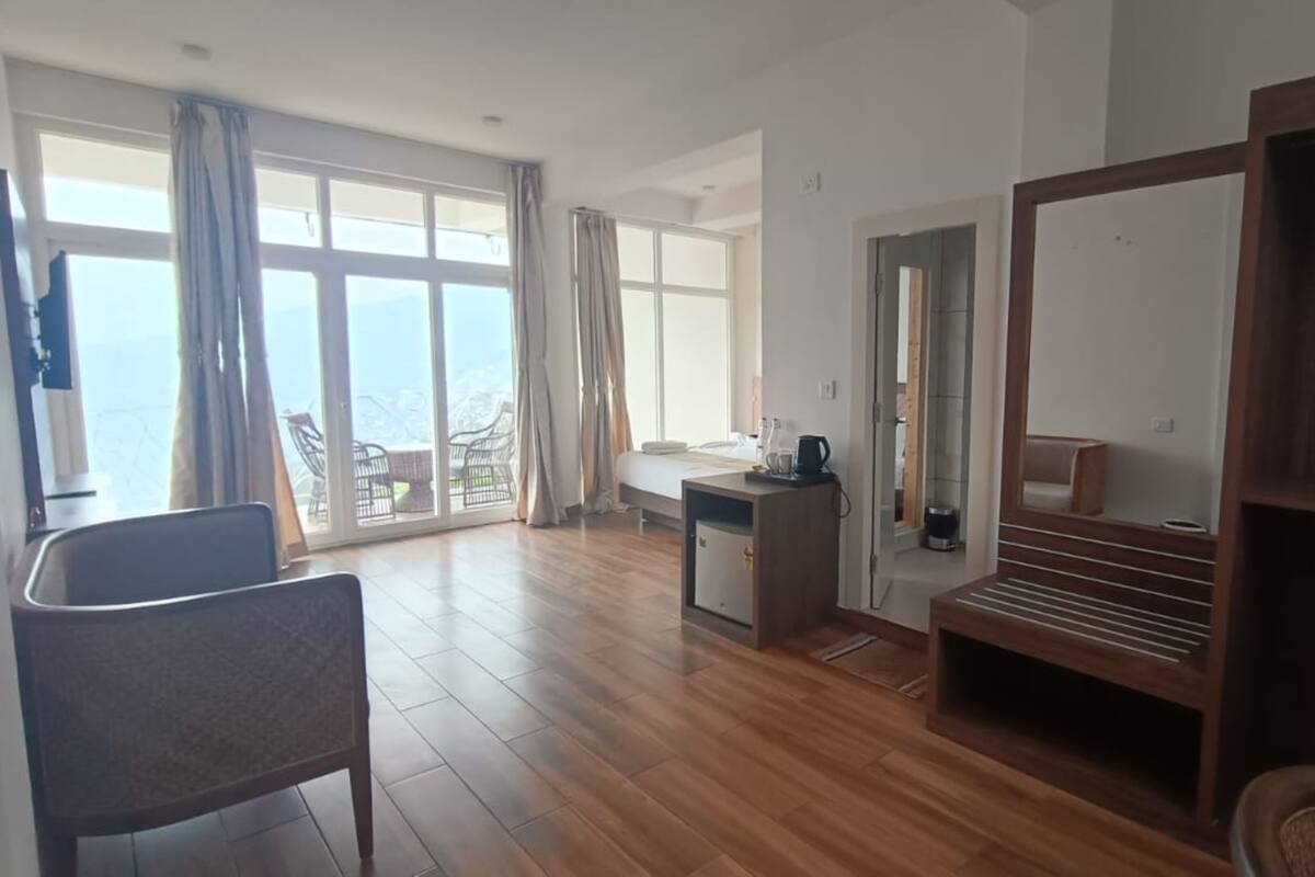 Een ruime kamer met houten vloer, een bed, bureau met stoel, en een groot raam met uitzicht op zee.