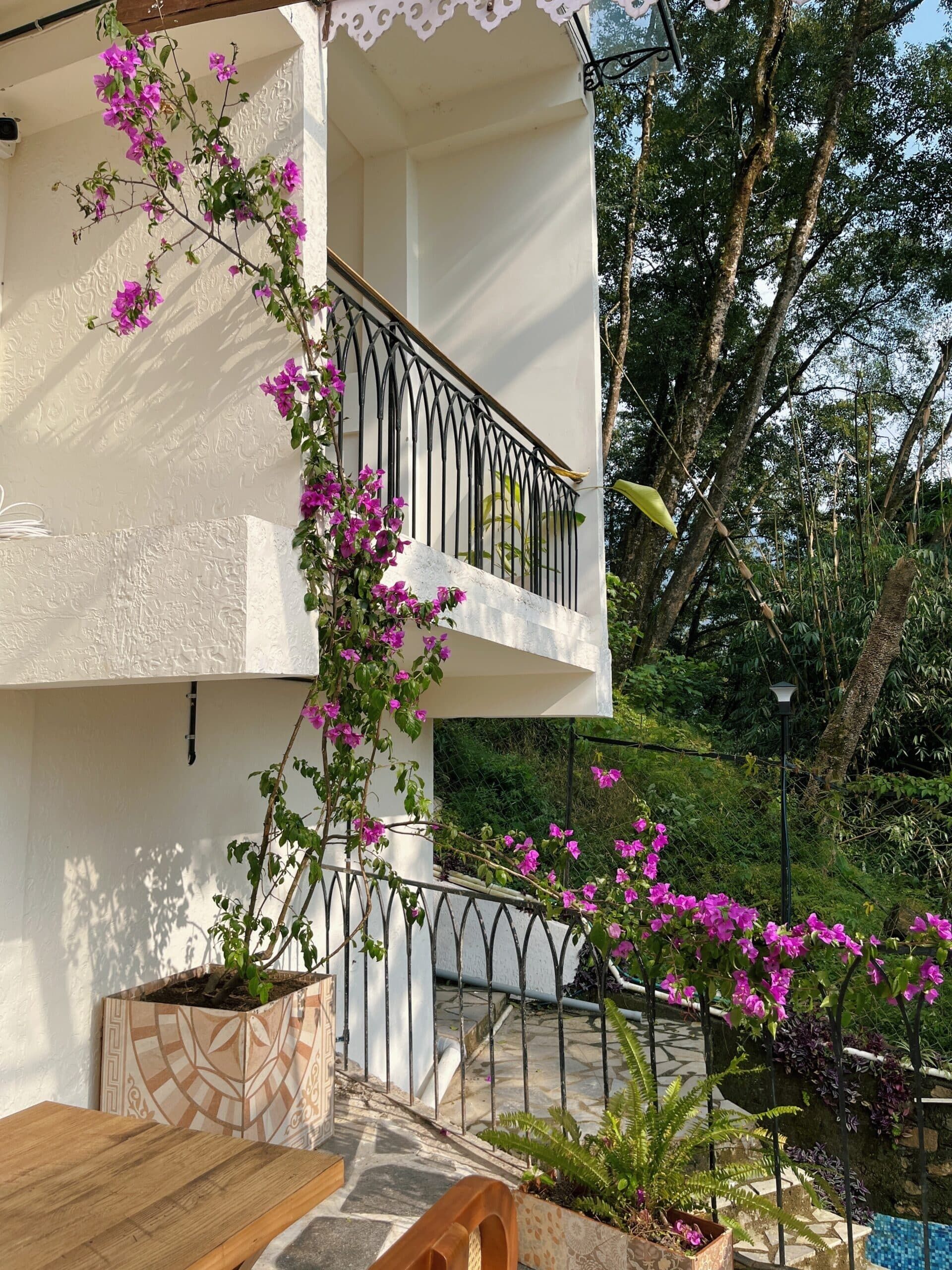 Un edificio de dos pisos con balcón, mobiliario exterior y un jardín lleno de flores rosas.