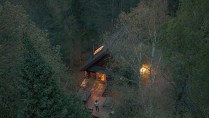 A cabin surrounded by dense forest, with a wooden deck and a person standing near the entrance.