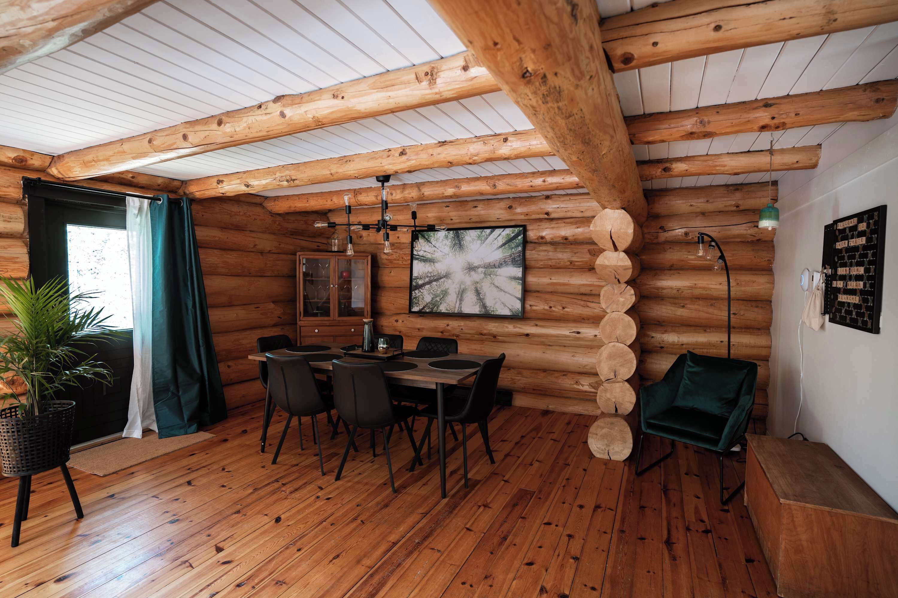 A rustic wooden interior with a dining area, a large window with curtains, and a wall-mounted artwork.