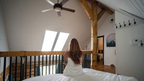 A room with a bed, a wooden ceiling, and a view of another room through a window.