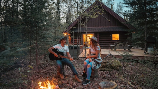 Two people sitting by a campfire in a forest, with a rustic cabin in the background.