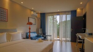 A hotel room with a large bed, a desk, a chair, a lamp, and a view through a glass door. - Harmony Hotel Kampala (Kampala)