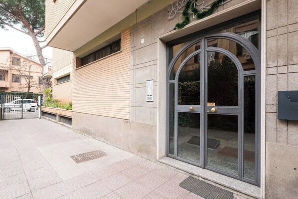 A building entrance with a large arched glass door, a small outdoor seating area, and a gated driveway. - Attico con vista Ciampino Roma  (Ciampino)