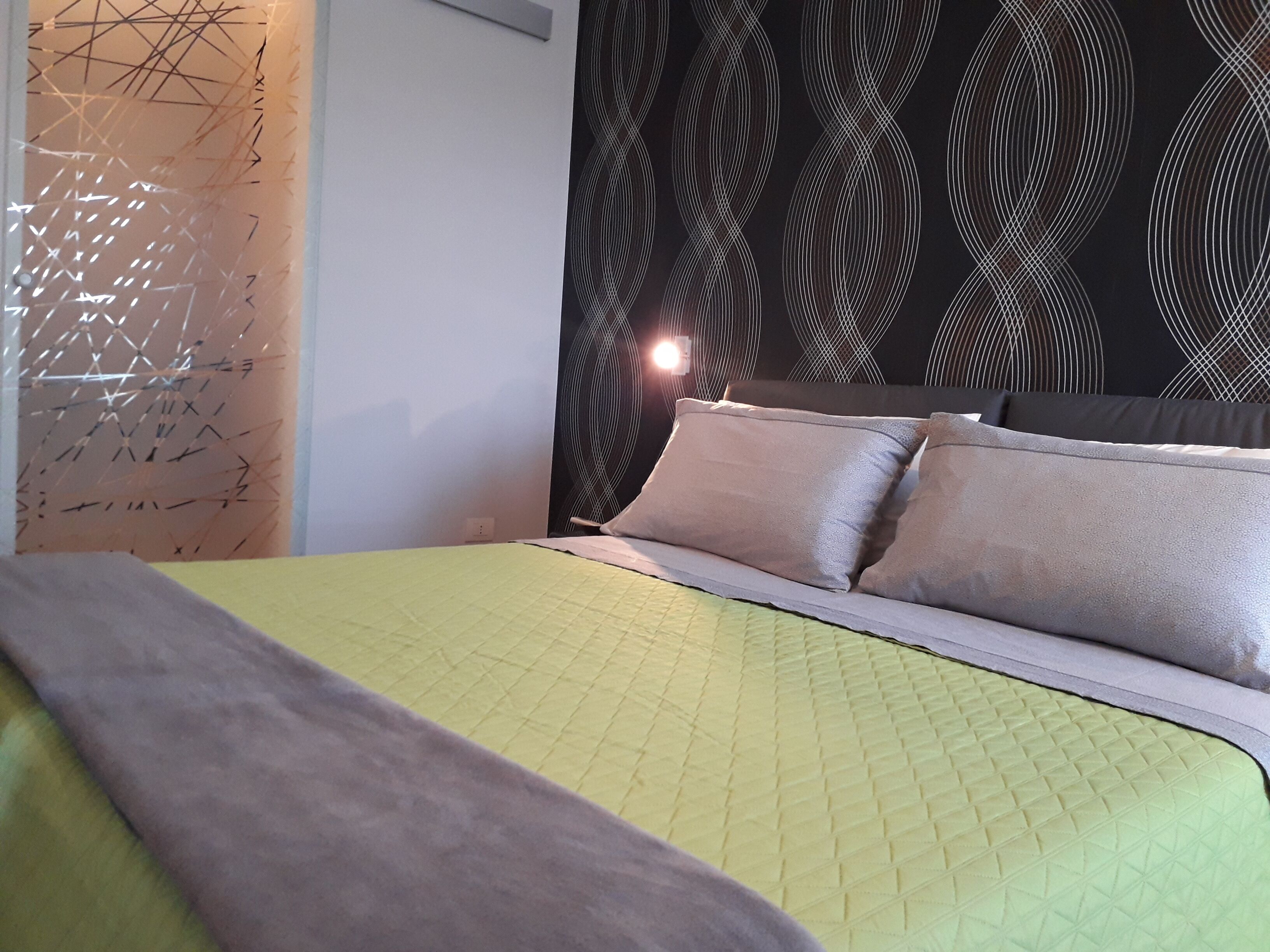 A bed with a quilted green bedspread and two pillows, a wall with a geometric pattern, and a headboard with a black and white design.