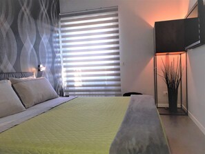 A bedroom with a bed, a window with blinds, a wall-mounted TV, and a floor lamp. - Attico con vista Ciampino Roma  (Ciampino)
